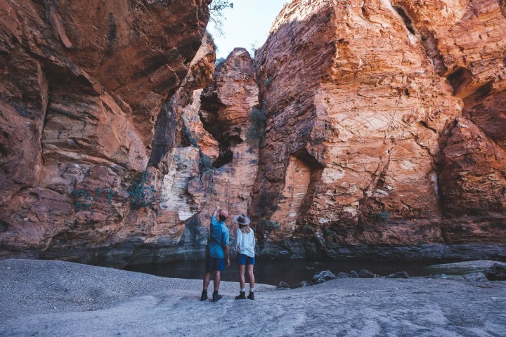 Two people stand by towering red rock formations with a clear blue sky.