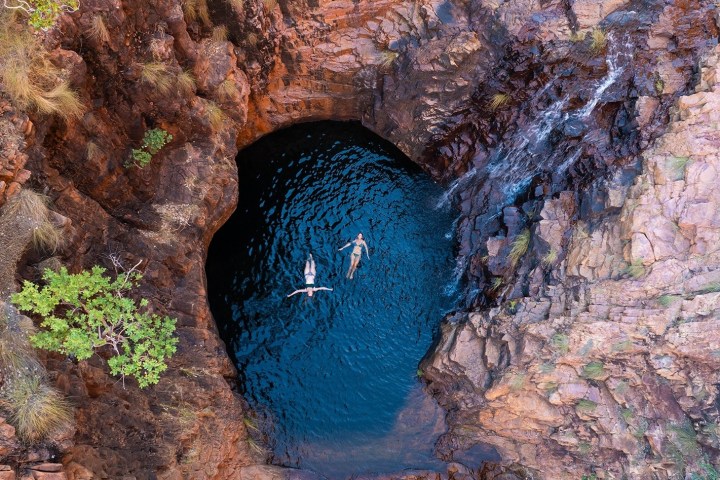 Two people swim in a natural rock pool surrounded by rugged cliffs.