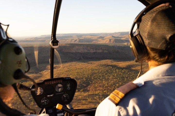 Two people in a helicopter cockpit flying over a desert landscape.