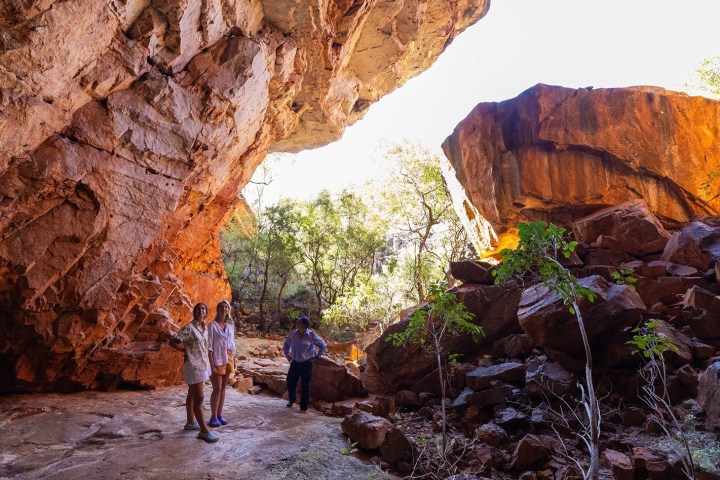 Three people stand under a large rocky overhang, surrounded by red rocks and greenery.