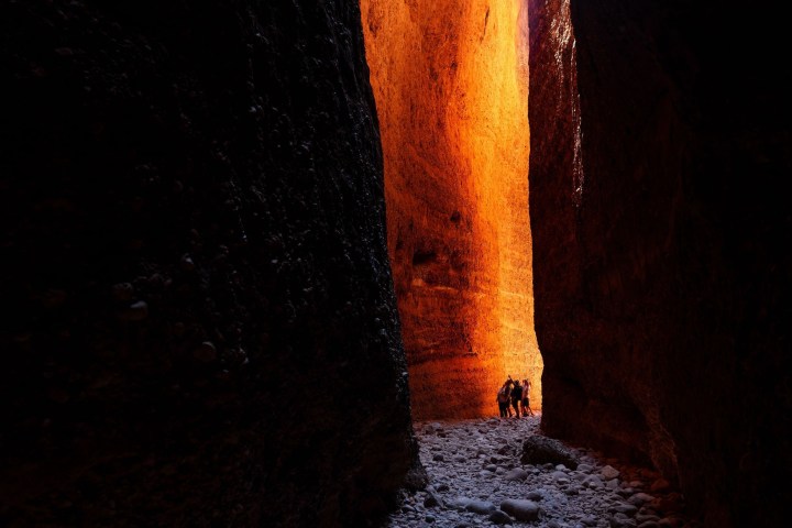 People walking in a narrow canyon with sunlight on orange rock walls.