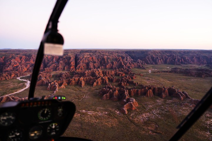 View from helicopter over reddish rock formations and green plains.