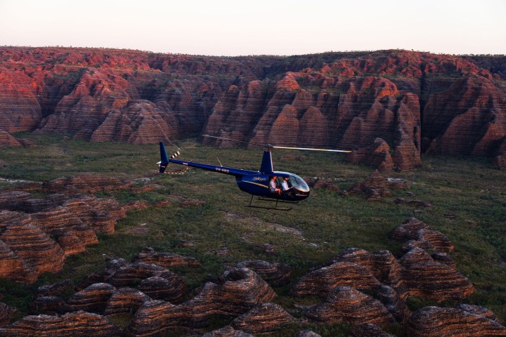 Blue helicopter flying over rugged, striped hills at sunset.