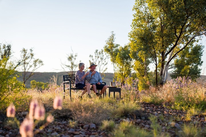 Two people sit on a bench in a grassy field with trees and flowers.