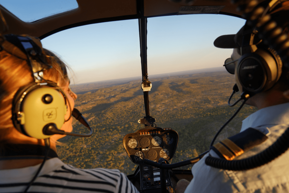 Two people in helicopter cockpit flying over a landscape at sunset.