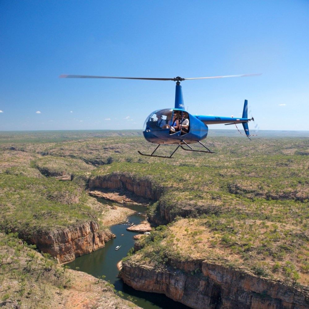 Blue helicopter flying over a canyon with a winding river and sparse vegetation.