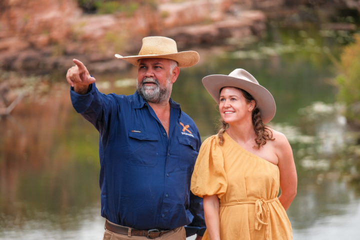 Man in hat points while woman in yellow dress smiles by water.