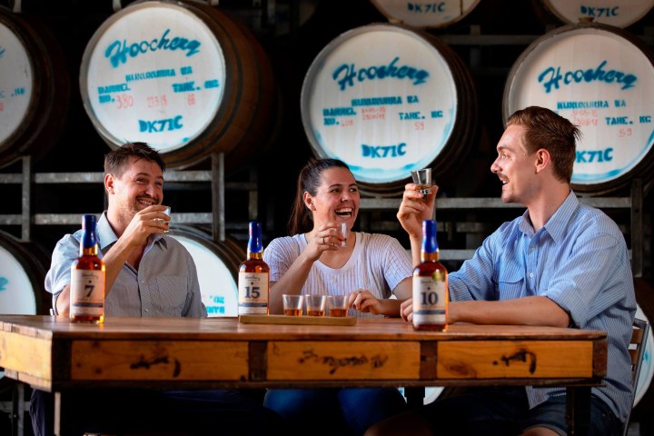 Three people enjoy whiskey at a table in front of barrels.
