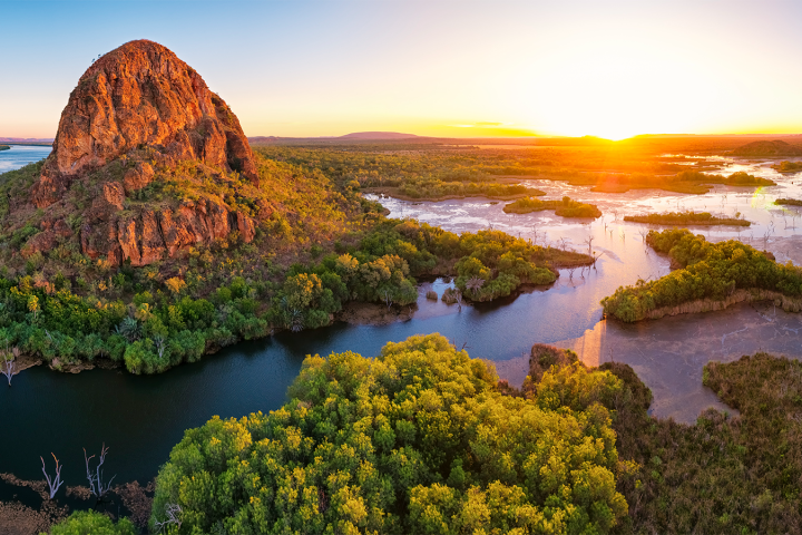 Aerial view of a large rock formation and river at sunrise, surrounded by lush green vegetation.
