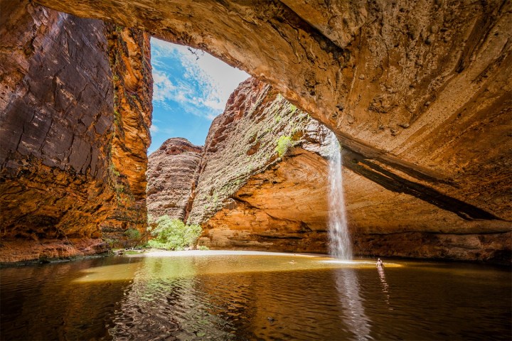 Cave with waterfall and pond, surrounded by rocky cliffs under a bright blue sky.