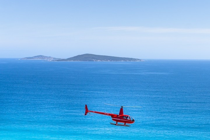 Red helicopter flying over blue ocean with distant island.