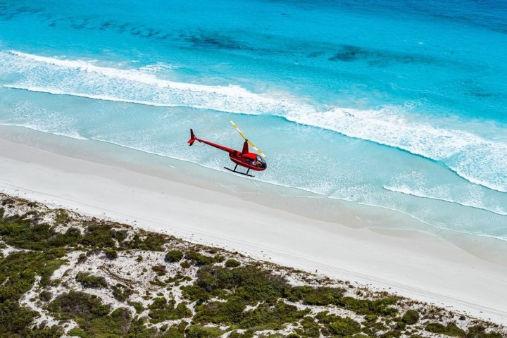 A red helicopter flying over a beach with turquoise water and white sand.