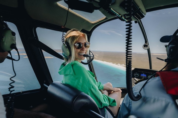 Three people inside a helicopter flying over a coastal landscape.