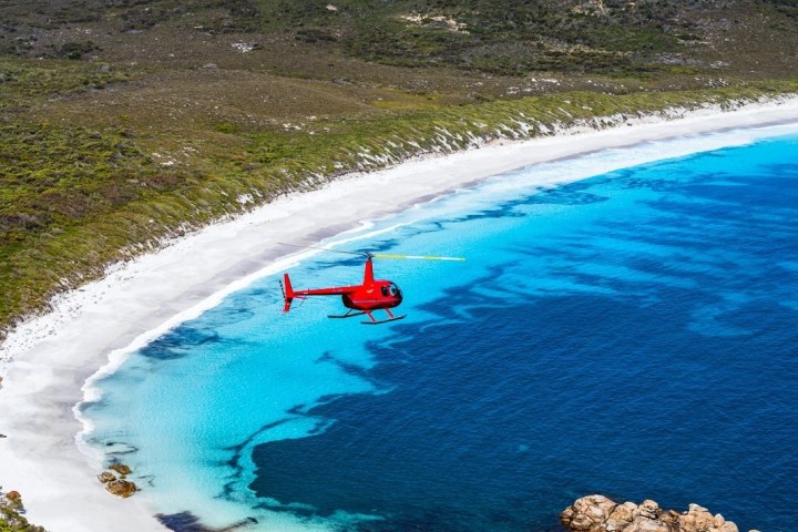 Red helicopter flying over white sandy beach and turquoise ocean.