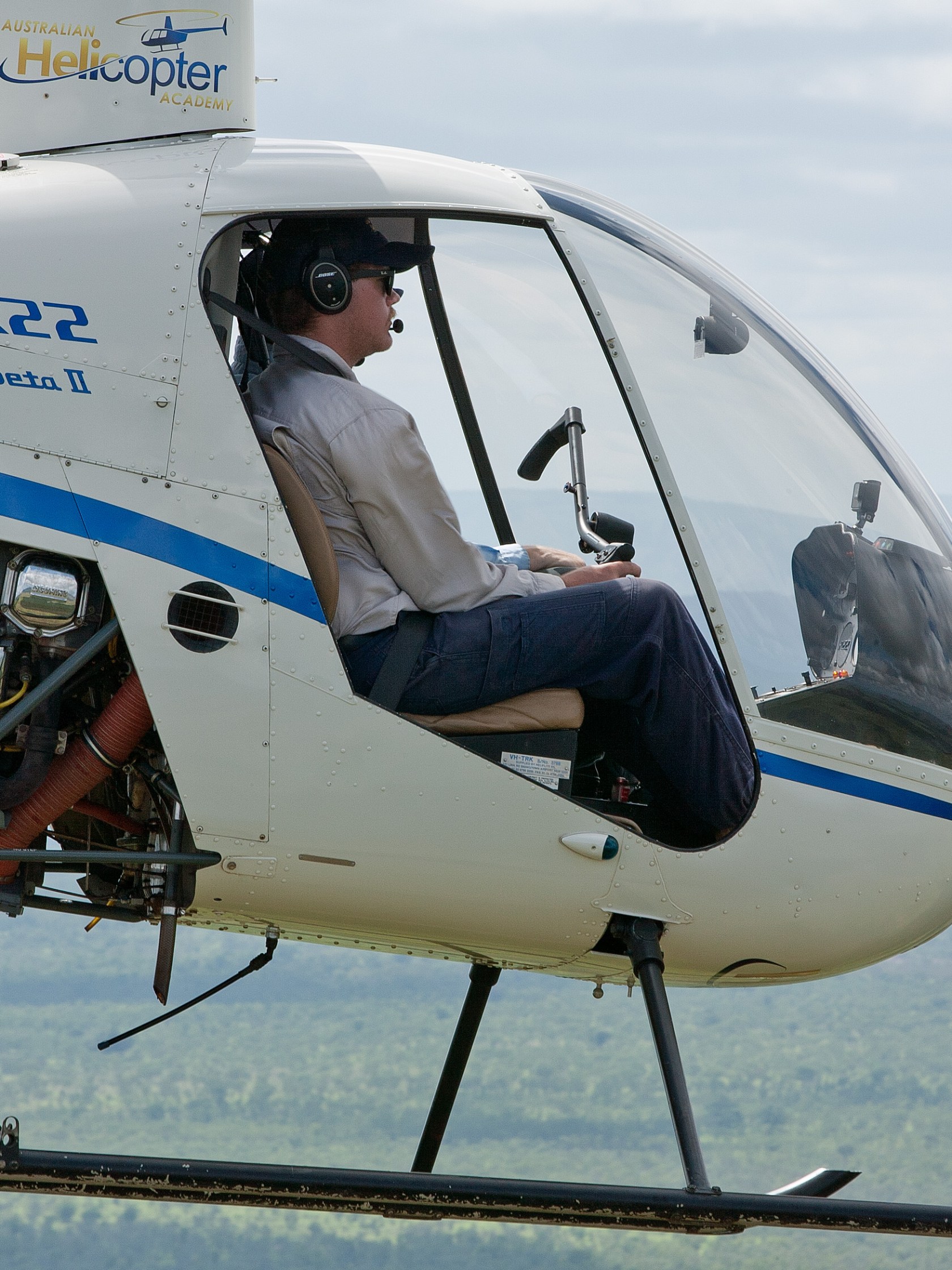 Man piloting small helicopter mid-flight over green landscape.