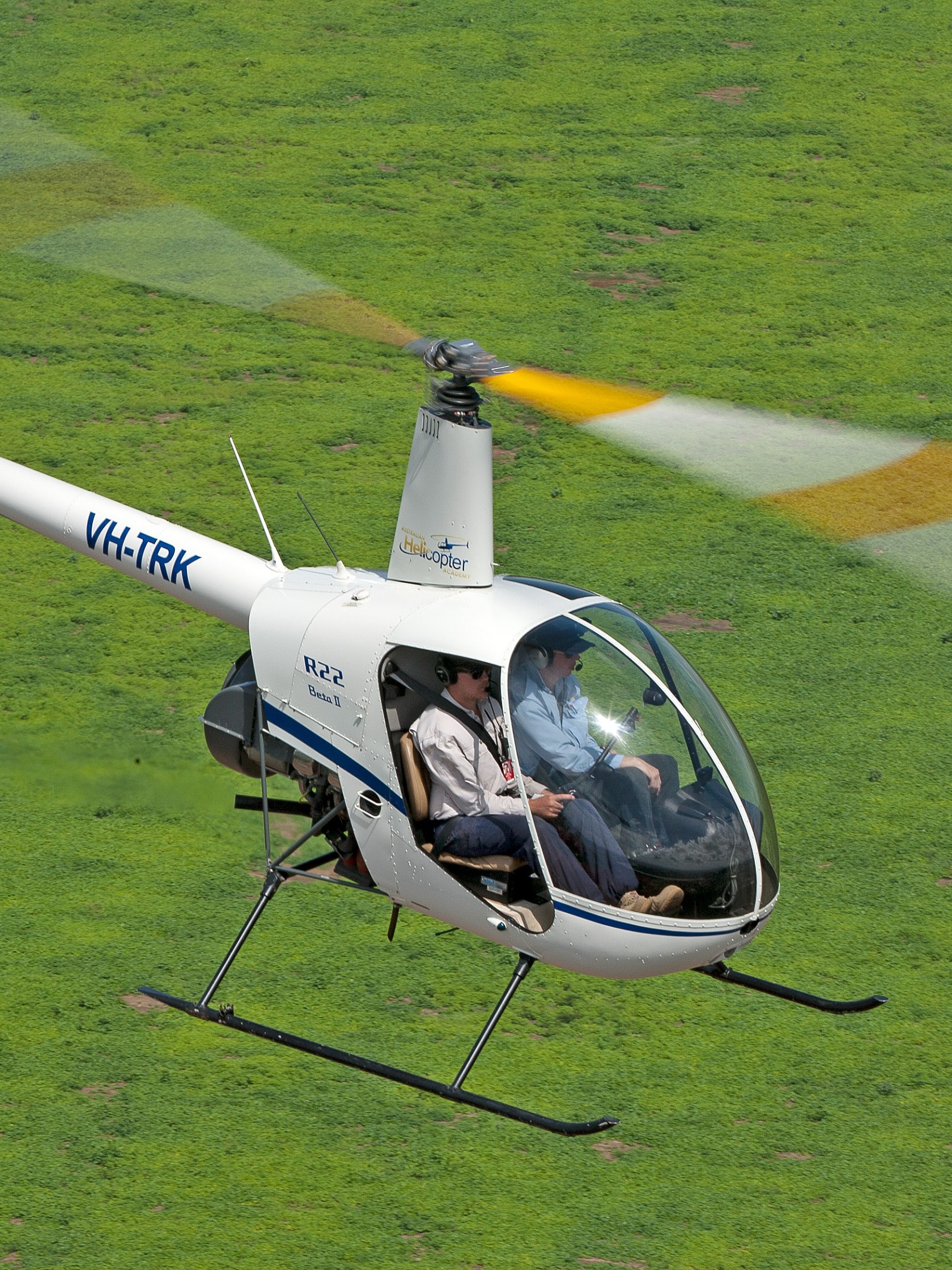 White helicopter with two passengers flying over green grass.