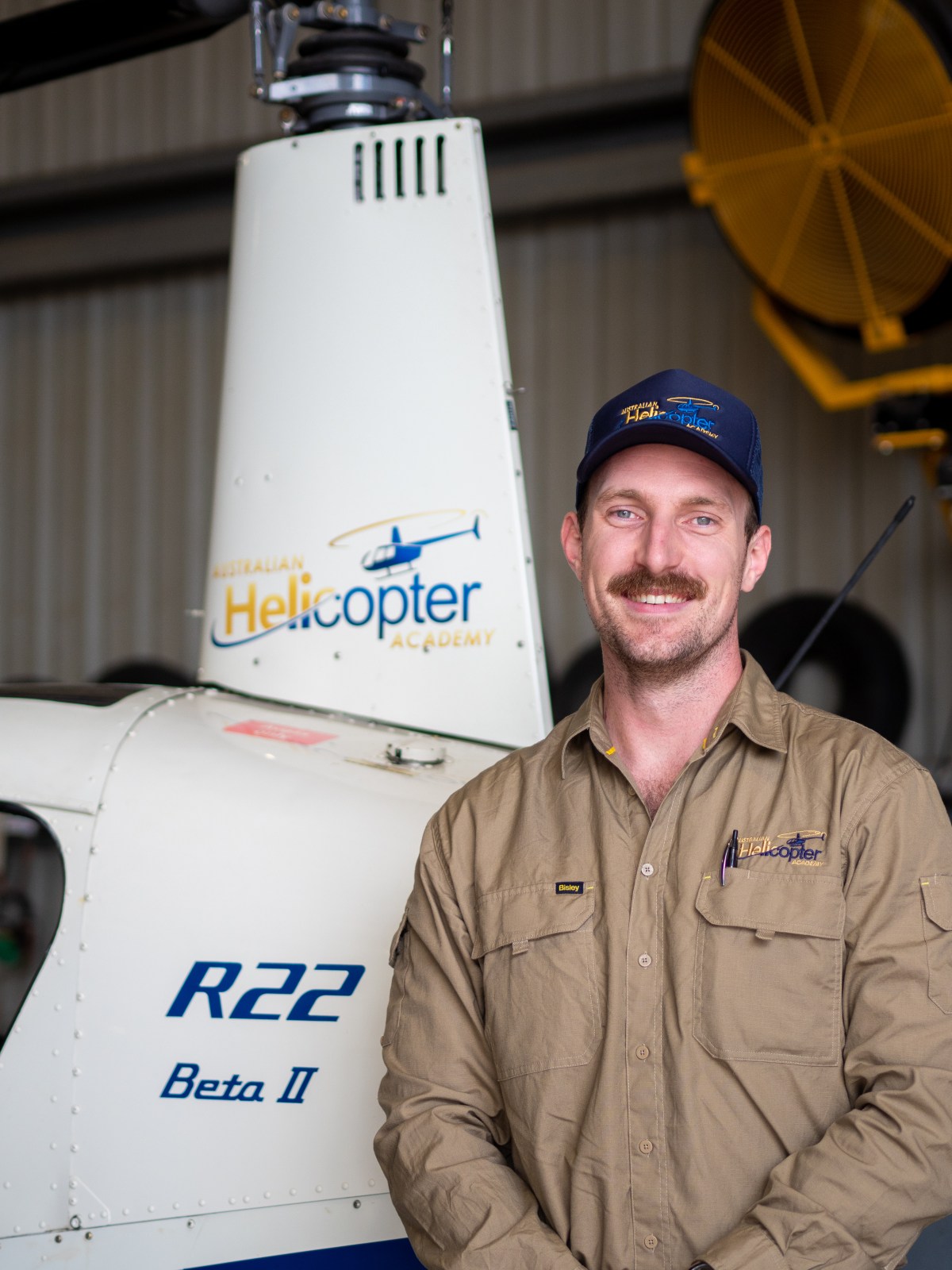 Smiling man in khaki shirt and cap stands by white helicopter inside a hangar.