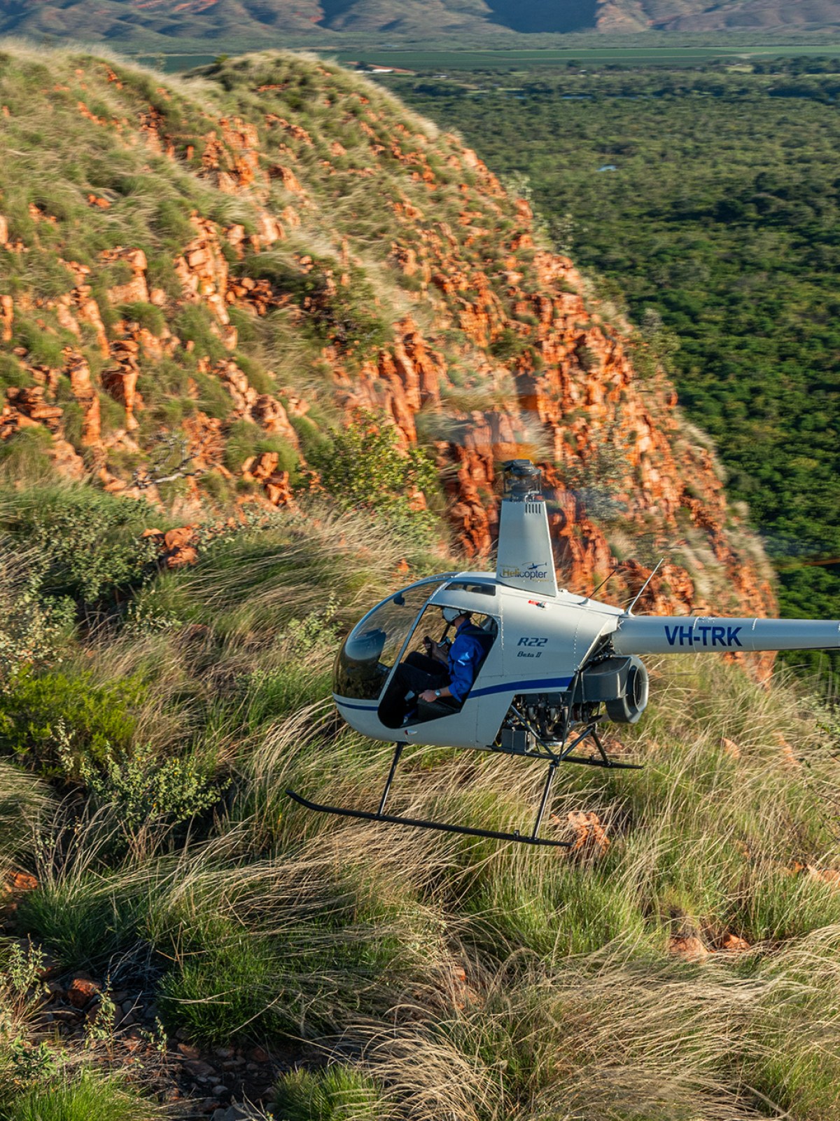 Helicopter flying over scenic, grassy hill with river and mountains in background.