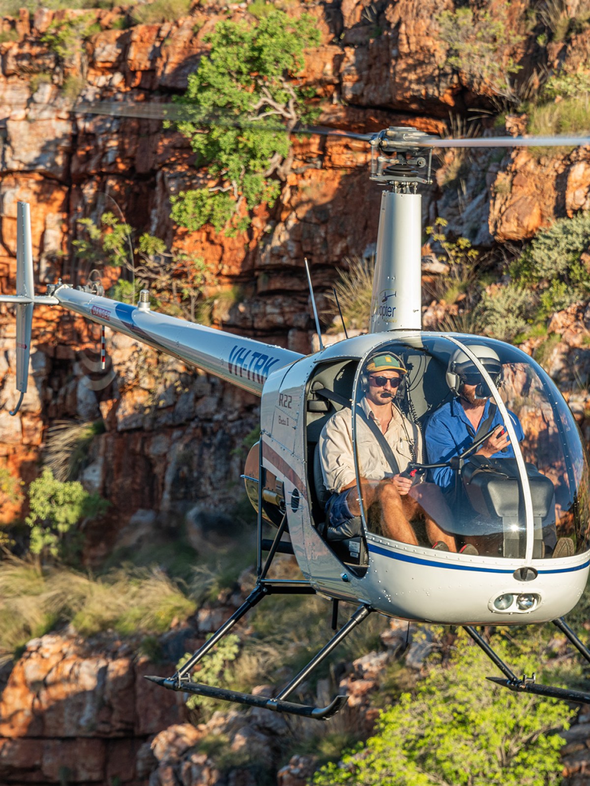 Helicopter flying near a rugged, rocky cliff with two people inside.