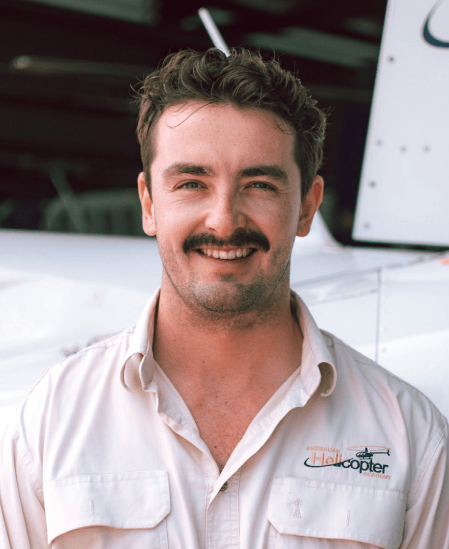 Man smiling with a mustache, wearing a light shirt, standing in front of an aircraft.