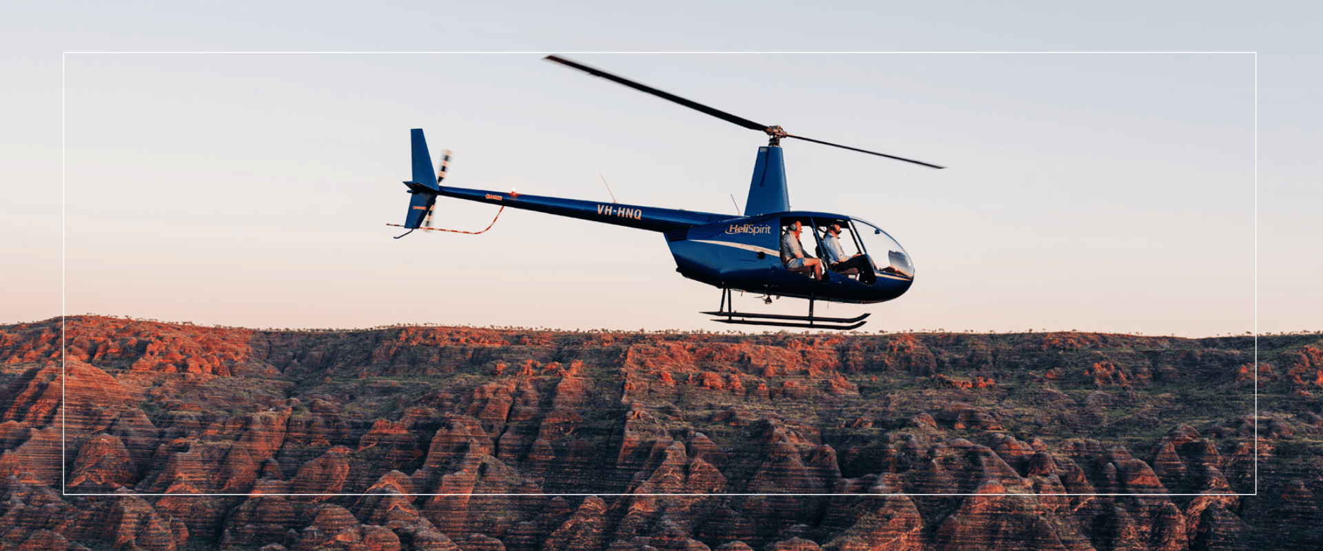 Blue helicopter flying over rocky terrain at sunset.