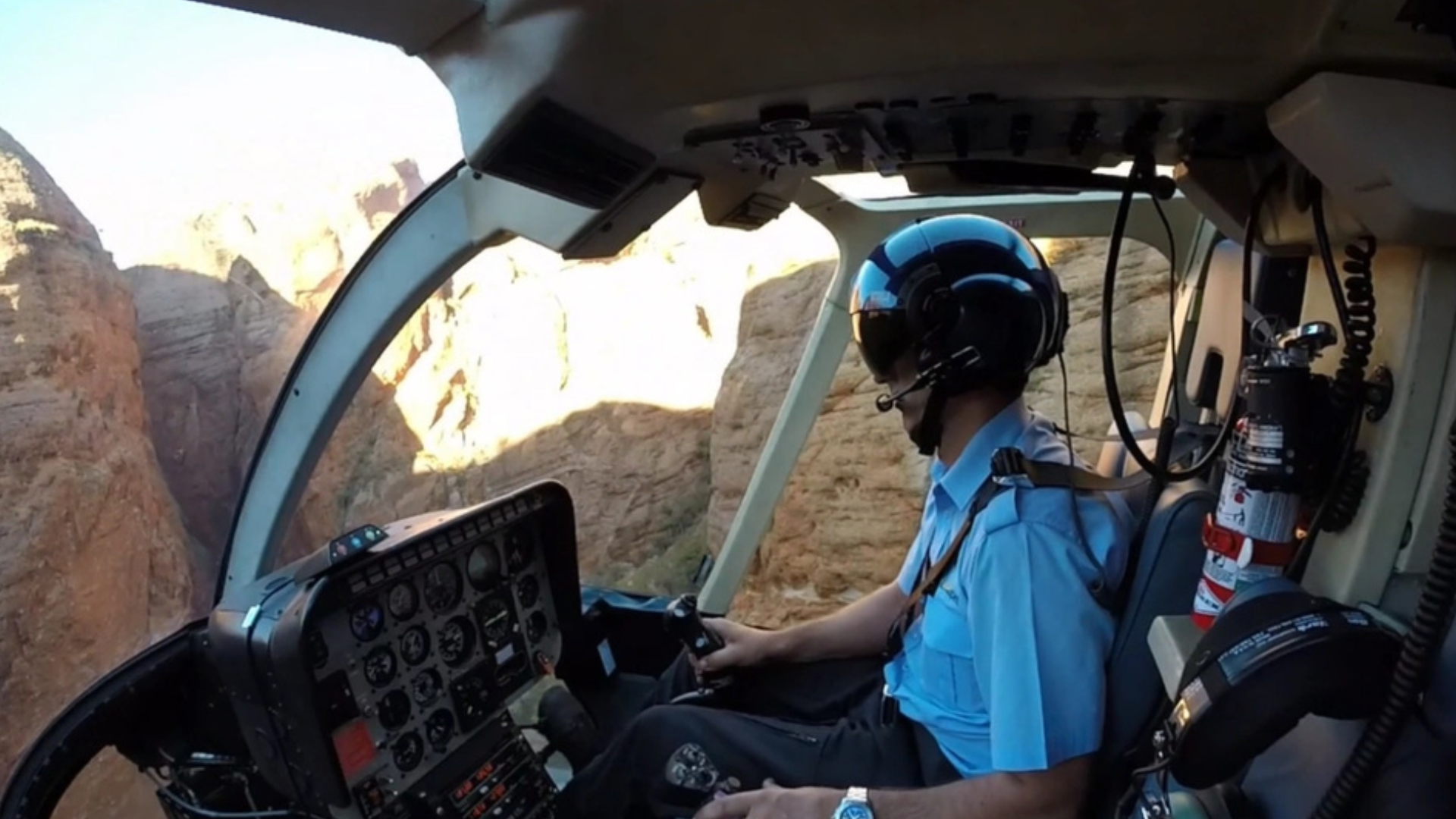 Pilot in cockpit flying a helicopter over rocky canyon landscape.