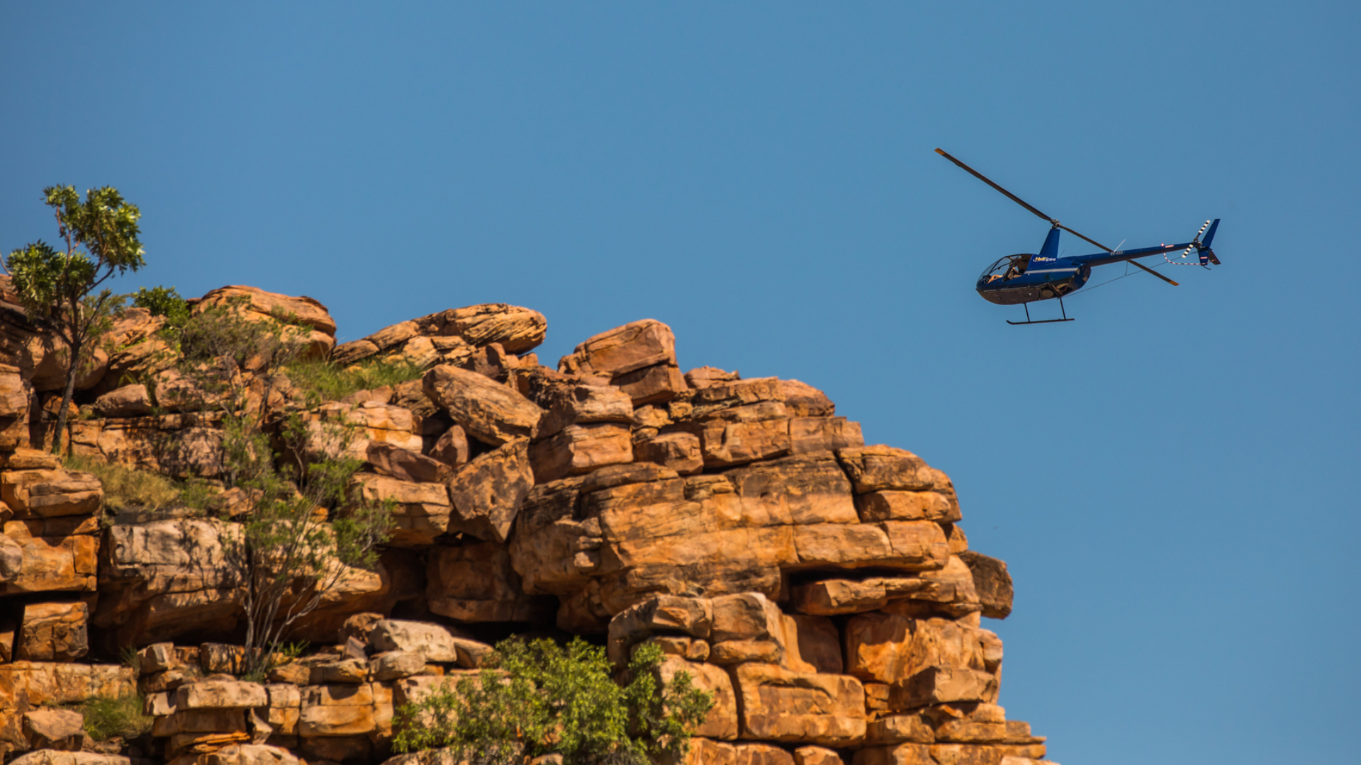 Helicopter flying over a rocky cliff under a clear blue sky.