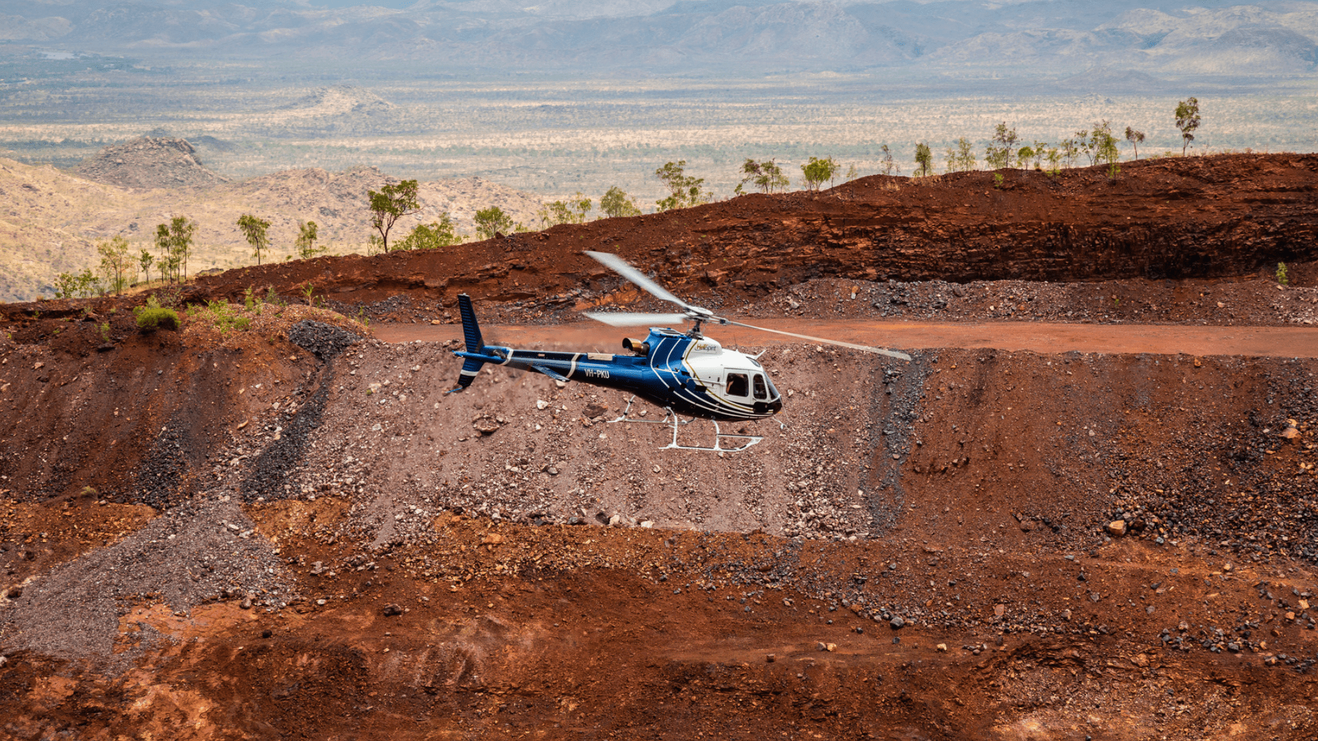 Blue helicopter flying over rocky, hilly landscape with sparse trees.