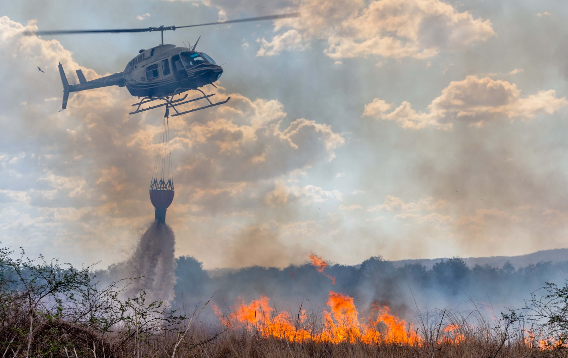 Helicopter dropping water on a wildfire in a grassy area with smoke and flames.