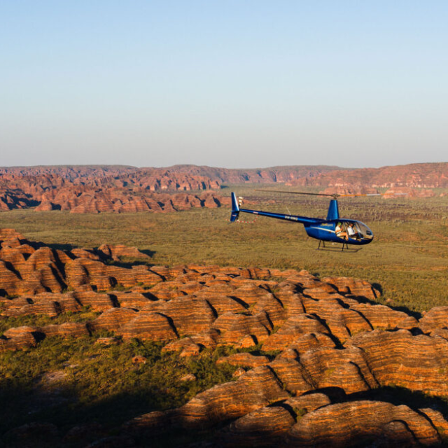 Blue helicopter flying over a rocky, desert landscape under a clear sky.