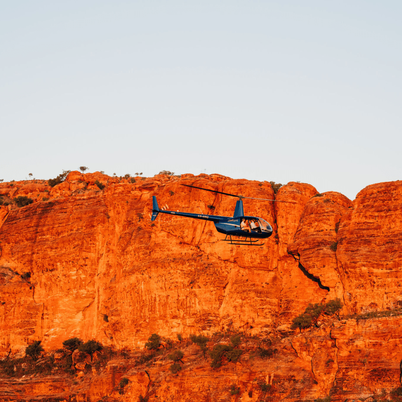 Blue helicopter flying near a red rocky cliff under clear sky.