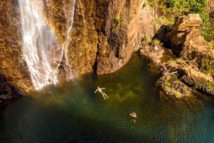 Two people swimming near a waterfall with a rainbow in a rocky area.